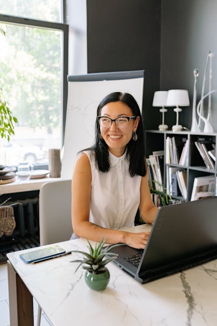 pexels-photo-8837175-8837175 Smiling Asian businesswoman using laptop in a stylish, well-lit office with plants.